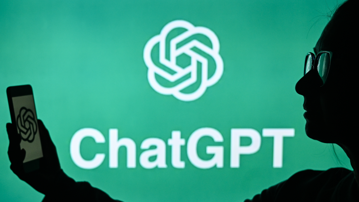 A woman holds a cell phone in front of a computer screen displaying the ChatGPT logo
