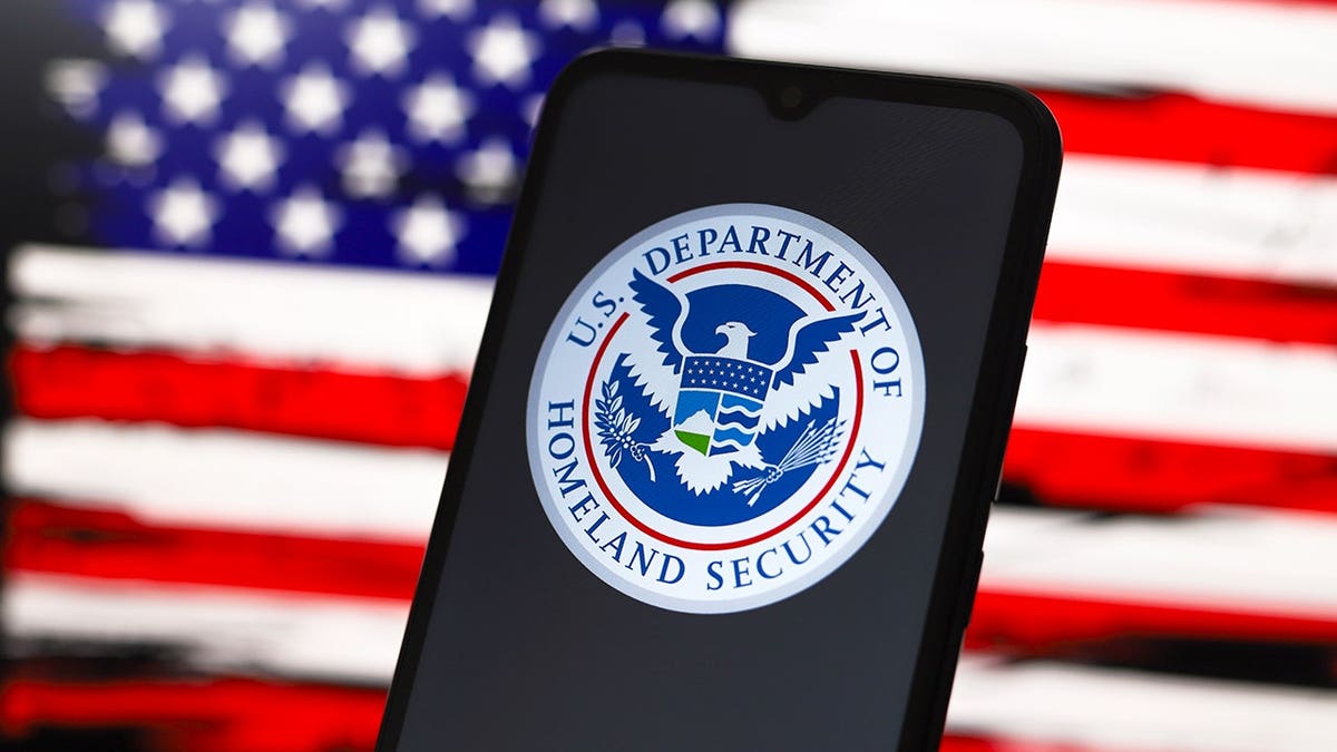 Department of Homeland Security logo on smartphone screen with American flag design on computer screen in background