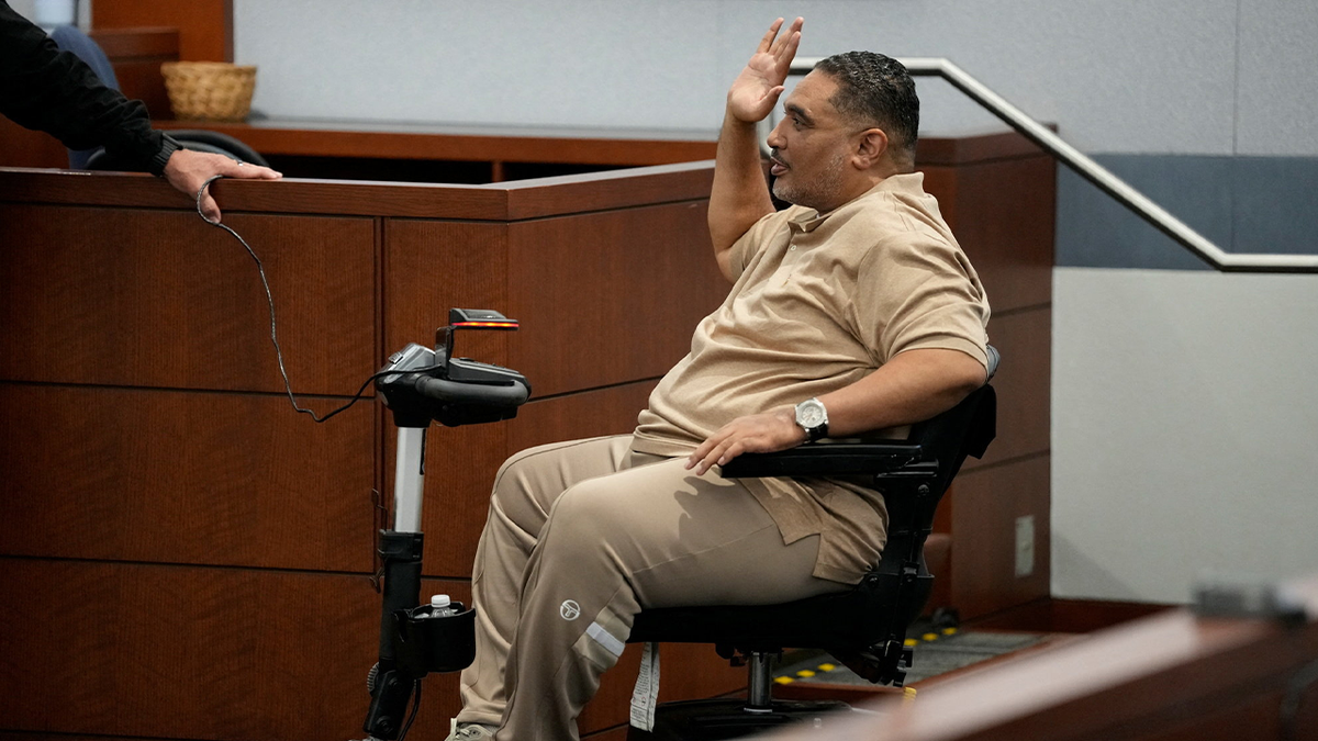 Reggie Wright Jr raises one hand to swear in before taking the witness stand, he is wearing a tan outfit and seated in a motorized wheelchair