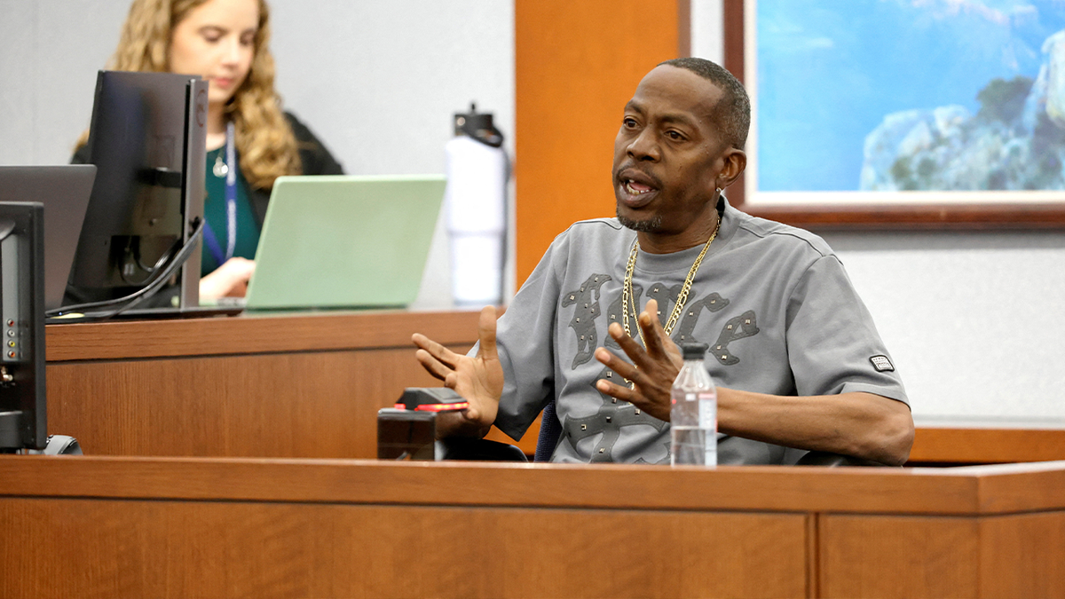 Denvonta Lee gestures from the witness stand with both hands, wearing a gray T-shirt and gold chain