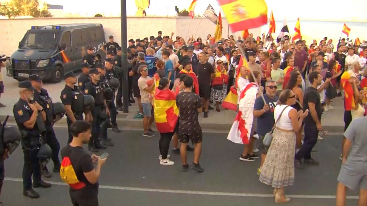 Law enforcement personnel watch while protesters hold Spanish flags, as Ceuta residents destroy migrant camp by the beach