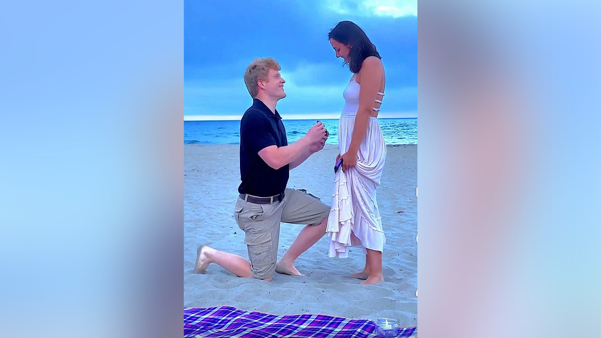 Cameron Lyon kneels on a beach while proposing to his fiancée, Abrielle Kerns
