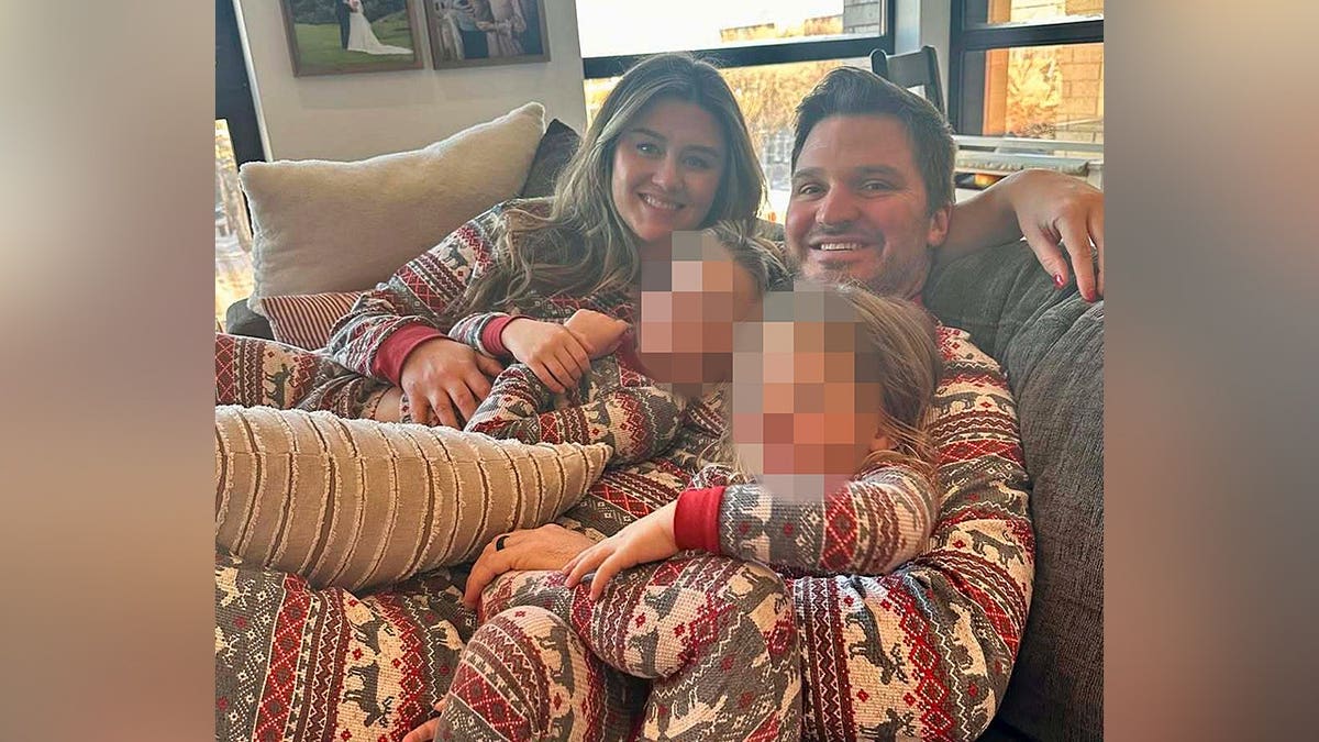 Corie Walsh and her husband smiling on a sofa wrapped around two young children wearing matching holiday pattern pajamas.