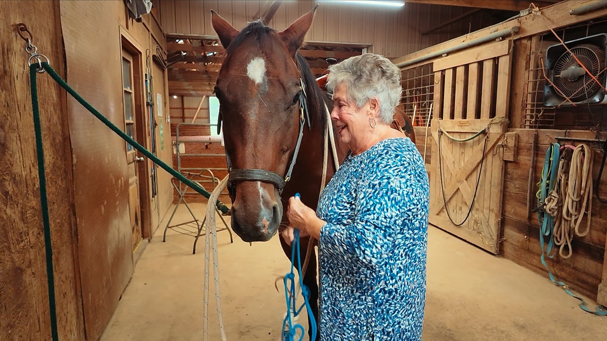 Horse ranch owner Lynette Ralph smiles beside a brown horse in a wooden stable.