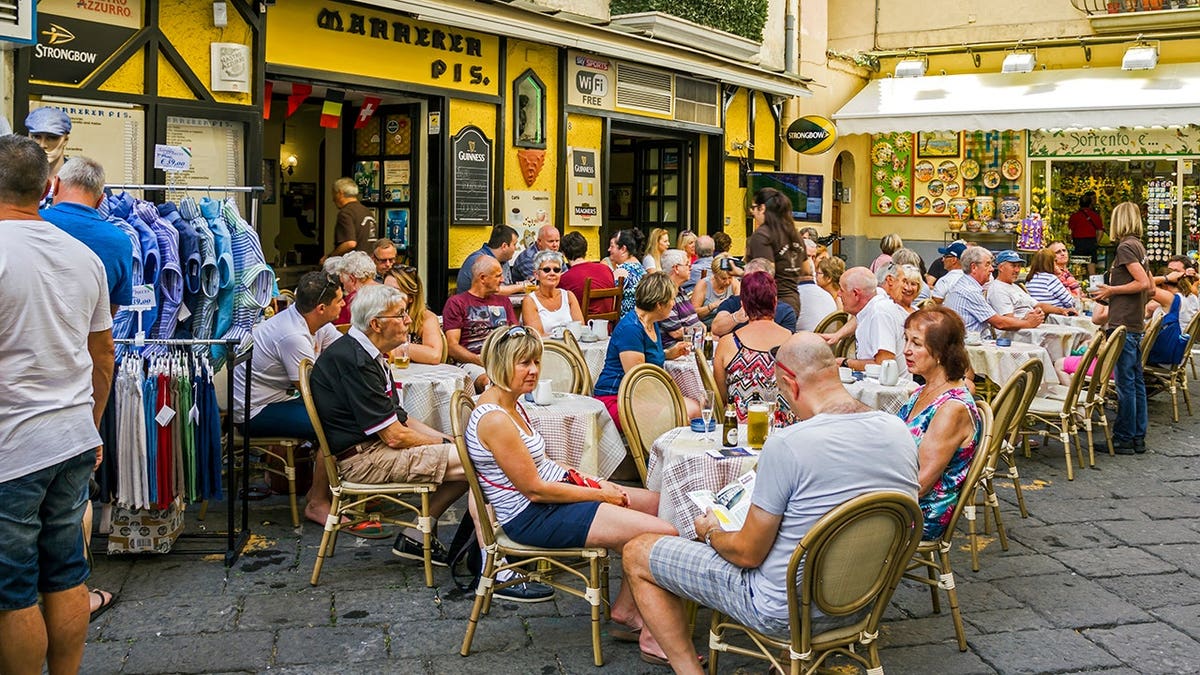 People sit at outdoor tables and walk along a promenade on a main street in Sorrento, Italy.